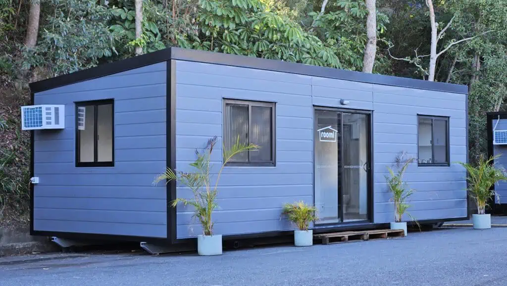 Single-room portable structure with horizontal blue-grey cladding and black window frames, designed as a relocatable living or working space